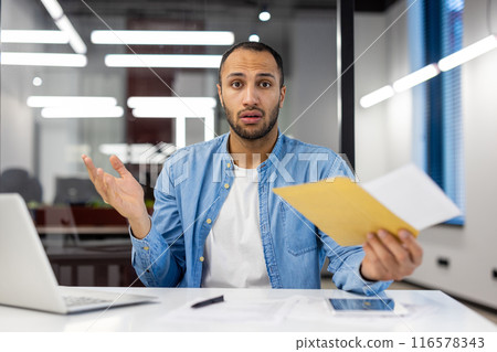 Portrait of upset and disappointed young hispanic man sitting at desk in office and received bad news by letter, looking worried at camera. 116578343