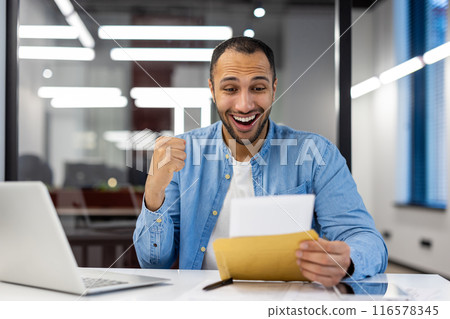 Smiling young hispanic man sitting in modern office and reading letter received in mail, happy with news and message. Smiling young hispanic man sitting in modern office and reading letter received in mail, happy with news and message. 116578345