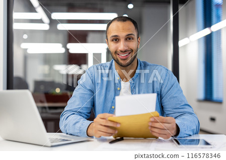 Portrait of a young hispanic man sitting at a desk in a modern office, holding an open letter in an envelope and smiling at the camera. Portrait of a young hispanic man sitting at a desk in a modern office, holding an open letter in an envelope and smiling at the camera. 116578346
