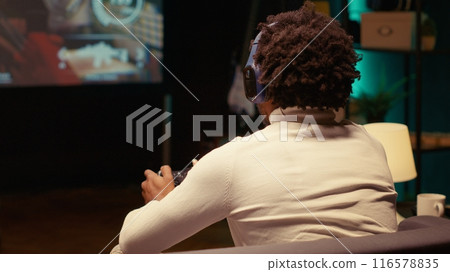 Close up shot of BIPOC man playing SF FPS videogame on widescreen smart TV, having fun in dark apartment room. Gamer competing in online multiplayer game, using headphones to chat with teammates Close up shot of BIPOC man playing SF FPS videogame on widescreen smart TV, having fun in dark apartment room. Gamer competing in online multiplayer game, using headphones to chat with teammates 116578835