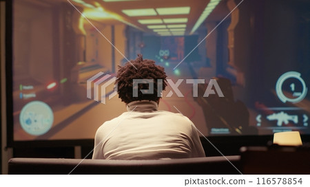 Man playing first person shooter videogame on smart TV in futuristic industrial setting with health, stamina, ammo bars HUD overlay. Gamer enjoying competitive PvP fps online multiplayer game Man playing first person shooter videogame on smart TV in futuristic industrial setting with health, stamina, ammo bars HUD overlay. Gamer enjoying competitive PvP fps online multiplayer game 116578854