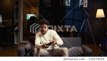 Man in neon lit home theatre playing video games on couch, enjoying day off from work. Gamer battling enemies in online multiplayer shooter on gaming console, using motion controlled controller Man in neon lit home theatre playing video games on couch, enjoying day off from work. Gamer battling enemies in online multiplayer shooter on gaming console, using motion controlled controller 116578856