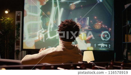 Man plays first person shooter videogame on widescreen TV with automatic gun shooting laser bullets at flying robotic foes. Gamer partakes in PvP online multiplayer game tourney 116578919