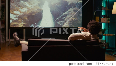 African american man watching nature documentary with beautiful cinematography on video projector. Person enjoying mountains and rivers scenery shots in home theatre, relaxing on couch African american man watching nature documentary with beautiful cinematography on video projector. Person enjoying mountains and rivers scenery shots in home theatre, relaxing on couch 116578924