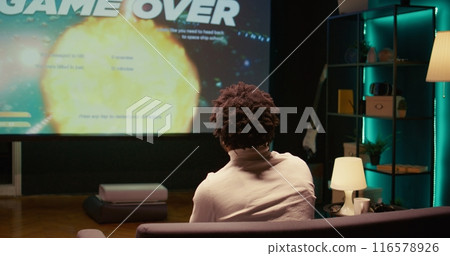 Upset african american gamer raging after losing game, playing on ultrawide smart TV. Man in living room feeling angry and shouting after his spaceship exploded, defeated in videogame Upset african american gamer raging after losing game, playing on ultrawide smart TV. Man in living room feeling angry and shouting after his spaceship exploded, defeated in videogame 116578926