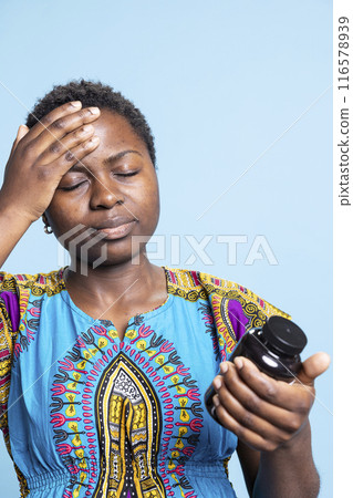 African american person feeling sick in front of the camera, reading label on medicine jar over blue background. Woman with headache feeling under pressure with serious illness, medicaments bottle. 116578939