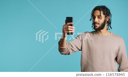 Joyous man greeting friend over online videocall on cellphone, doing friendly gestures. Excited person bonding with mate using mobile phone videoconference, studio background, camera A Joyous man greeting friend over online videocall on cellphone, doing friendly gestures. Excited person bonding with mate using mobile phone videoconference, studio background, camera A 116578945