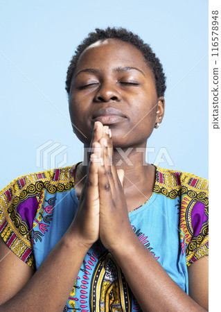 Confident serious person holding hands in a prayer to talk to God, saying grace and interacting with Jesus Christ in studio. Religious pious woman praying to the Lord, african american clothing. 116578948