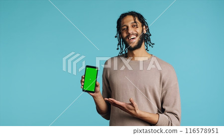 Happy man pointing towards green screen mobile phone, showing thumbs up sign. Cheerful person presenting chroma key cellphone, doing positive gesturing, isolated over studio background, camera A 116578951