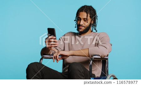 Man in wheelchair chatting with friend over internet videocall using smartphone. Person with disability chatting with mate in videoconference online meeting over blue studio background, camera B Man in wheelchair chatting with friend over internet videocall using smartphone. Person with disability chatting with mate in videoconference online meeting over blue studio background, camera B 116578977