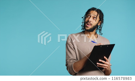 Middle Eastern photographer holding clipboard in hand, completing shoot list for upcoming photography work. Creative man thinking of ideas, writing on paper using pen, studio background, camera A 116578983