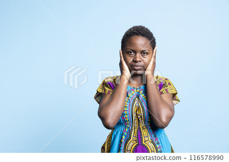 African american woman covering her ears in front of the camera, creating three wise monkeys concept sign. Female model presenting wisdom and iconic metaphor over blue background. 116578990