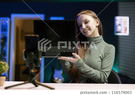 Happy gen Z online show host filming content about best laptop releases of the year with smartphone on tripod. Upbeat young influencer recording newly launched notebook review using mobile phone 116579003