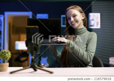 Joyous teenager presenting laptop from brand sponsoring video, urging subscribers to purchase it, filming with mobile phone. Upbeat girl does influencer marketing, recording with cellphone on tripod Joyous teenager presenting laptop from brand sponsoring video, urging subscribers to purchase it, filming with mobile phone. Upbeat girl does influencer marketing, recording with cellphone on tripod 116579016