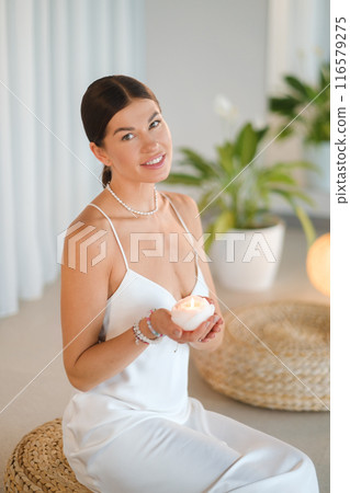 Beautiful Woman in a white dress with a candle in her hands sitting indoors before meditation Beautiful Woman in a white dress with a candle in her hands sitting indoors before meditation 116579275