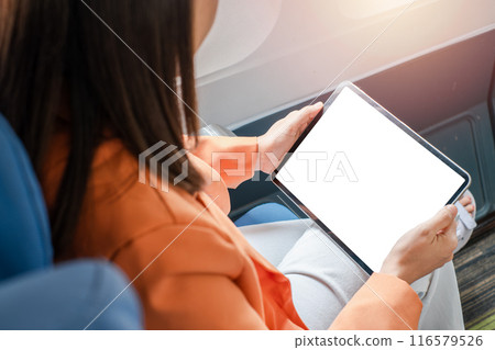 A woman in an orange jacket using a digital tablet with a blank screen while seated on an airplane, ideal for travel, technology, and business themes. 116579526