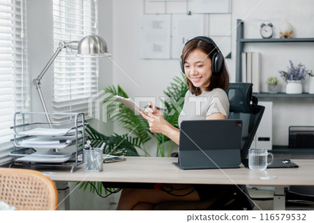 A young woman wearing headphones, working from home office with a tablet, taking notes, and smiling. A young woman wearing headphones, working from home office with a tablet, taking notes, and smiling. 116579532
