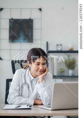 A female doctor in a white coat working on a laptop in a modern office setting, surrounded by medical equipment and documents. A female doctor in a white coat working on a laptop in a modern office setting, surrounded by medical equipment and documents. 116579543