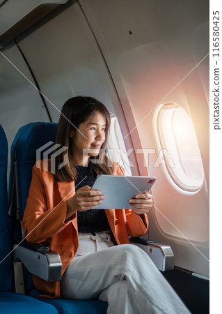 A young woman in a bright orange blazer uses a tablet while seated on an airplane, enjoying in-flight entertainment during her travel. 116580425