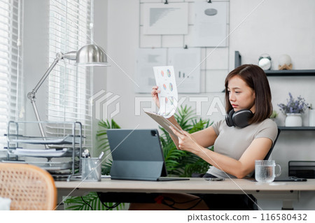 A young professional woman reviews business charts in a modern home office setting, equipped with a laptop and headphones. A young professional woman reviews business charts in a modern home office setting, equipped with a laptop and headphones. 116580432