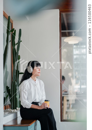 A young woman wearing headphones and holding a coffee cup, sitting in a modern minimalist cafe with cactus decor. 116580463