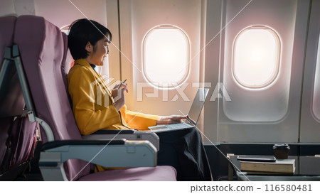 Professional woman in yellow jacket using laptop on airplane, working during flight, seated by window with coffee and book. 116580481