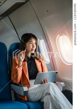 Professional woman in business attire working on a tablet while seated in an airplane, gazing thoughtfully out the window. 116580490