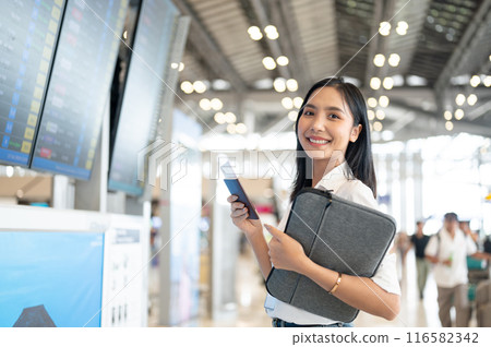 A beautiful Asian woman with her passport is standing in front of a flight information display. 116582342