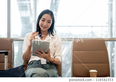 A charming Asian female passenger is working remotely, using her digital tablet in the airport. A charming Asian female passenger is working remotely, using her digital tablet in the airport. 116582353