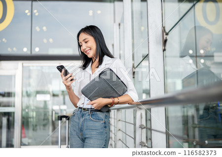An Asian female passenger is using her smartphone while standing at the exit door of the terminal. An Asian female passenger is using her smartphone while standing at the exit door of the terminal. 116582373