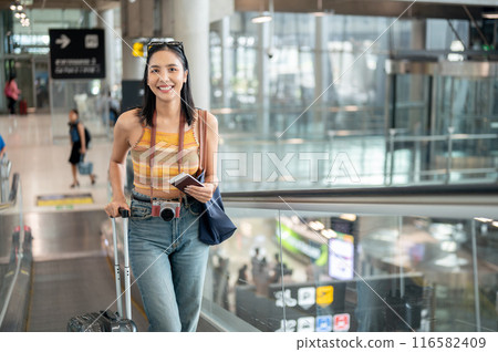 A beautiful Asian female tourist with her luggage is on the airport escalator, traveling by plane. A beautiful Asian female tourist with her luggage is on the airport escalator, traveling by plane. 116582409