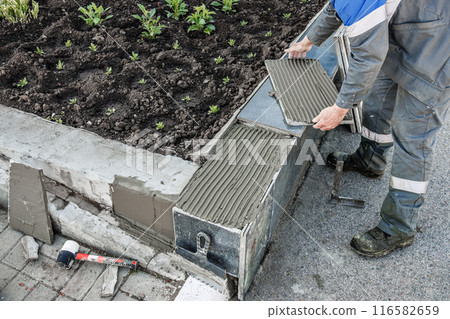 A man is laying bricks on a sidewalk. The sidewalk is made of concrete and has a brick border. The 116582659