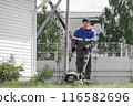 A man in a blue and gray jacket is mowing the lawn. He is wearing a hat and gloves 116582696