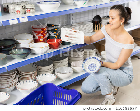 Positive Asian woman choosing plates in dishware store 116583657
