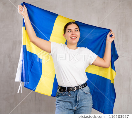 Joyous young woman holding Sweden flag against unicoloured background Joyous young woman holding Sweden flag against unicoloured background 116583957