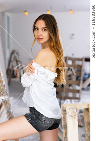 Long-haired half-naked woman in shirt dropped from shoulders covering breasts with hands posing in room under renovation 116583960