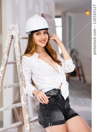 Brown-haired woman in construction hardhat, shorts and unbuttoned blouse standing in apartment during overhaul Brown-haired woman in construction hardhat, shorts and unbuttoned blouse standing in apartment during overhaul 116584127