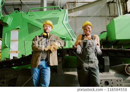 Medium long low angle view shot of African American woman and Caucasian man standing relaxed against industrial machine at work in factory Medium long low angle view shot of African American woman and Caucasian man standing relaxed against industrial machine at work in factory 116584558