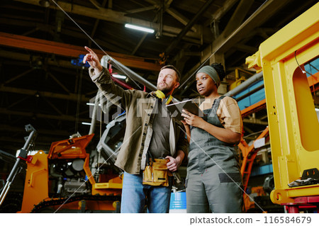 Low angle view of mature Caucasian engineer showing new equipment to his Black female colleague at work in modern factory 116584679