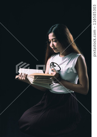 Young Asian woman with long hair kneeing reading a book in low light, wearing white shirt and maroon skirt, holding rosary Young Asian woman with long hair kneeing reading a book in low light, wearing white shirt and maroon skirt, holding rosary 116585303