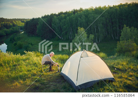 Extreme long shot of experienced senior male tourist sitting by camp tent cooking dinner, copy space 116585864
