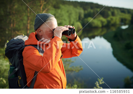 Medium shot of unrecognizable mature male tourist wearing backpack standing at bank of river looking through binoculars Medium shot of unrecognizable mature male tourist wearing backpack standing at bank of river looking through binoculars 116585931