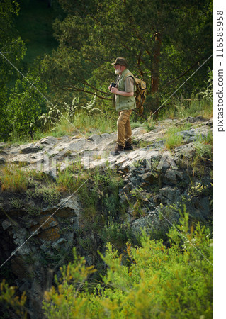 Vertical extreme long shot of senior Caucasian naturalist holding binoculars spending summer day in mountains, copy space 116585958