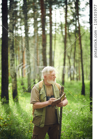 Vertical shot of senior Caucasian scientist working in mountain forest on summer day writing notes in notebook 116585977