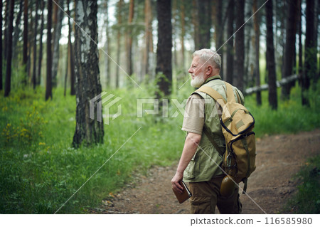 Medium long shot of senior male traveler wearing backpack holding notebook standing on forest road and looking away 116585980