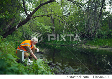 Wide shot of senior Caucasian male ecologist in safety vest taking samples at bank of river in forest on summer day, copy space 116586005