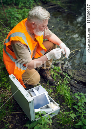 Vertical high angle view of senior male ecologist wearing safety vest with gloves taking river water sample 116586017