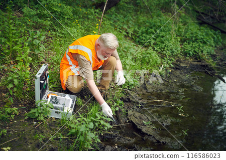 High angle view of senior scientist wearing orange safety vest taking water sample from river with pipette, copy space High angle view of senior scientist wearing orange safety vest taking water sample from river with pipette, copy space 116586023