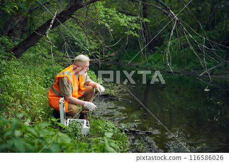 Senior Caucasian ecologist wearing orange safety vest and gloves sitting at bank or forest river taking water sample Senior Caucasian ecologist wearing orange safety vest and gloves sitting at bank or forest river taking water sample 116586026