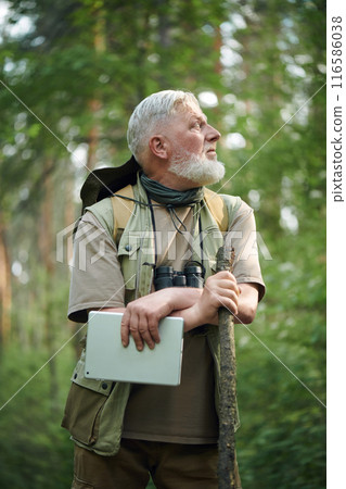 Vertical medium shot of pensive senior Caucasian botanist holding digital tablet standing in forest looking away Vertical medium shot of pensive senior Caucasian botanist holding digital tablet standing in forest looking away 116586038